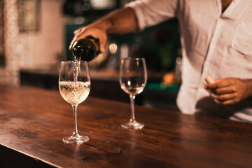 A barman pouring white wine into a glass. Wooden table in a dark bar
