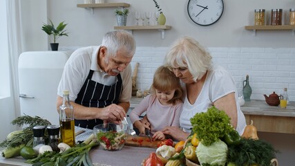 Senior couple in kitchen teaching granddaughter child how to cook, chopping pepper with knife