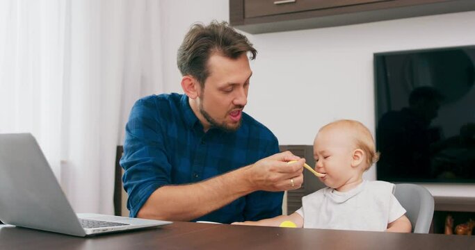 Young bearded father, and a cute baby with bib, in high baby chair, are sitting at the desk with laptop on it. Father feeds baby with bowl and spoon. Baby makes funny faces and grimaces. Slow motion