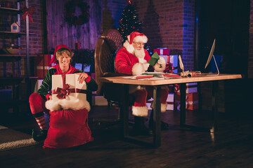Photo portrait of santa claus reading green letter from child and elf putting wrapped present into...