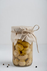 Single glass jar full of homemade organic marinated or fermented champignon mushrooms decorated with craft paper on white wooden background with black pepper at kitchen. Vertical orientation image