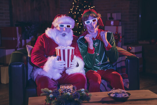 Photo portrait of shocked santa claus and elf holding popcorn watching movie on sofa in 3d glasses
