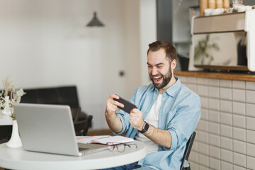 Fototapeta premium Cheerful man sit at table in coffee shop cafe working studying on laptop pc computer play game on cell phone relaxing in restaurant during free time indoor. Freelance mobile office business concept.