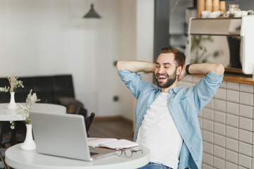 Obraz premium Cheerful young man sitting alone at table in coffee shop cafe restaurant indoors working or studying on laptop pc computer relaxing holding hands behind head. Freelance mobile office business concept.