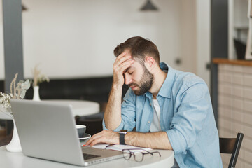 Naklejka premium Sick tired exhausted young man sitting alone at table in coffee shop cafe restaurant indoor working or studying on laptop pc computer put hand on head. Freelance mobile office business concept.