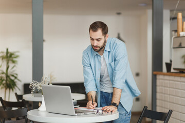 Handsome serious young man standing alone near table in coffee shop cafe restaurant indoors working or studying on laptop pc computer writing in notebook. Freelance mobile office business concept.
