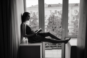 Pensive dreamy female holding book at home looking at big window dreaming, thoughtful young woman