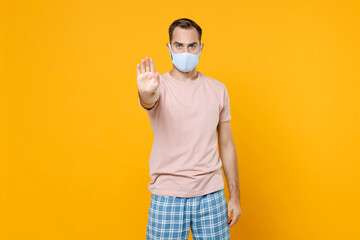 Fototapeta na wymiar Dissatisfied young man in pajamas home wear face mask safe from coronavirus virus covid-19 showing stop gesture with palm resting at home isolated on bright yellow background. Relax good mood concept.