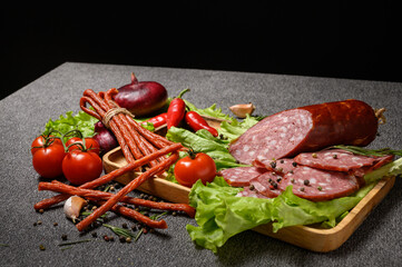 The slices sausage served on lettuce leaves in a wooden plate. Also thin long sticks of smoked sausage tied with string. Chili pepper, tomatoes on a sprig, onion and garlic. 