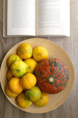 Wooden bowl with tangerines and a pumpkin and open book on a table. Top view.