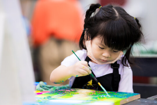 Cute girl is coloring on a canvas. Child holding paintbrush, paint and make art in classroom. Happy little children open the cloth mask from his mouth and nose. Kid are 3 years old