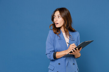 Shocked amazed beautiful attractive young brunette woman 20s wearing basic jacket hold clipboard with papers document writing looking aside isolated on bright blue colour background studio portrait.