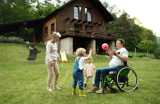 Small children with senior grandparents in wheelchair playing with a ball in garden.