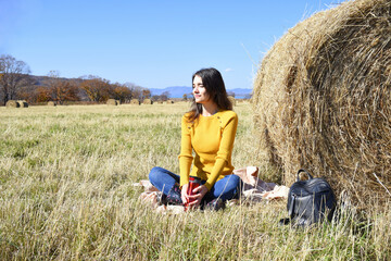 Young Russian brunette woman in yellow sweater and rubber boots sitting on plaid next to backpack near bale of hay in field. she holding thermos with tea,  from the sun and smiling. 
