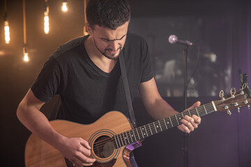 A young man in a black T-shirt plays the guitar at a concert. Blurry black background and warm...