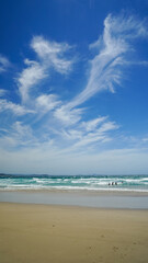 Windy day at the beach: featuring a blue sky with windswept clouds, people in the surf, and a sandy beach.