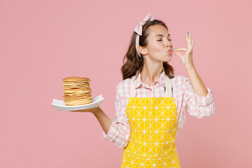 Young brunette woman housewife in yellow apron hold plate with pancakes making okay taste delight...