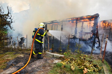 Firefighter uses water from hose to extinguish a huge fire of wooden structure, building, damage,...