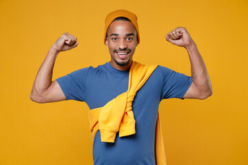 Smiling funny strong young african american man 20s wearing basic casual blue t-shirt hat standing showing biceps muscles spreading hands isolated on bright yellow colour background, studio portrait.