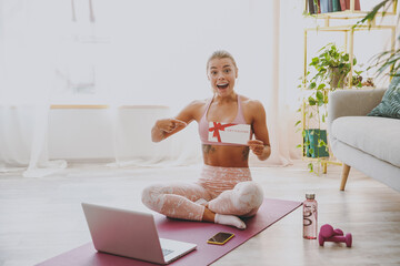 Surprised young strong sporty fitness woman wearing pink tracksuit doing yoga exercises using laptop computer pointing index finger on gift certificate sitting stretching on mat floor at home indoor.