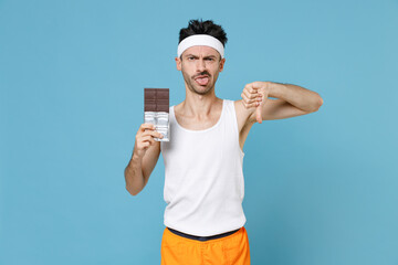 Dissatisfied young fitness man with thin skinny body sportsman in white headband shirt shorts hold chocolate bar showing thumb down isolated on blue background. Workout gym sport motivation concept.