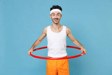 Excited young strong sporty fitness man with thin skinny body sportsman in white headband shirt shorts training with hula hoop isolated on blue background studio. Workout gym sport motivation concept. © ViDi Studio