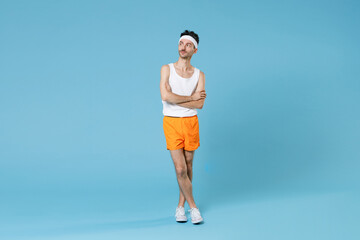 Fototapeta na wymiar Full length portrait of young sporty fitness man with skinny body sportsman in white headband shirt shorts holding hands crossed isolated on blue background. Workout gym sport motivation concept.