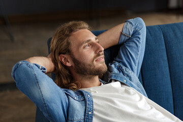 Fototapeta premium Time to relax. A curly-haired guy in a casual style, sitting on the couch at home, smiling and relaxing. Pleasant leisure time. Rest at home.