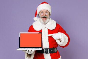 Excited Santa Claus man in Christmas hat red coat pointing index finger on laptop pc computer with blank empty screen isolated on violet background. Happy New Year celebration merry holiday concept.