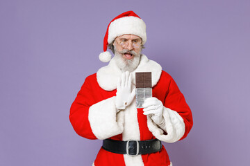 Fototapeta na wymiar Displeased Santa Claus man in Christmas hat red coat gloves glasses showing stop gesture with palm to chocolate bar isolated on violet background. Happy New Year celebration merry holiday concept.