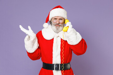 Confused Santa Claus man in Christmas hat red suit coat gloves glasses talking on telephone spreading hands isolated on violet background studio. Happy New Year celebration merry holiday concept.