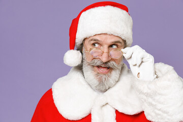 Excited Santa Claus man wearing Christmas hat red suit coat glasses keeping mouth open looking aside isolated on violet purple background studio. Happy New Year celebration merry holiday concept.