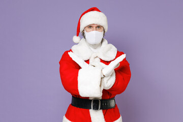 Fototapeta na wymiar Santa Claus man in Christmas hat suit coat mask safe from coronavirus virus covid-19 showing stop gesture with crossed hands isolated on violet background. New Year celebration merry holiday concept.