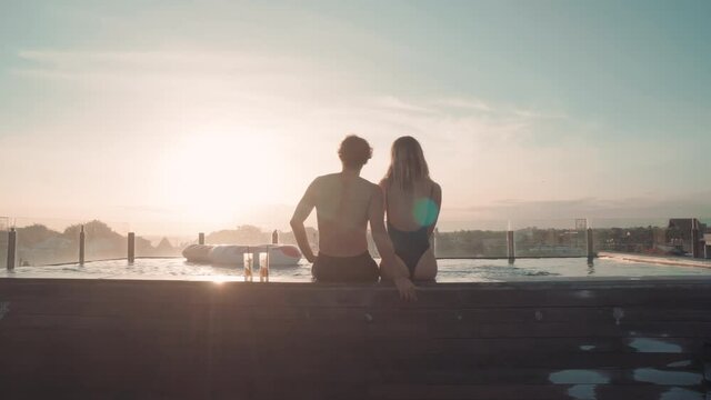 Dollying-out back-view of two lovers looking at sun under clear blue sky at outdoor terrace with swimming pool