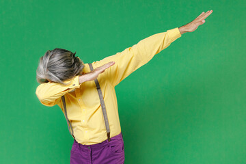 Elderly gray-haired mustache bearded man wearing casual yellow shirt suspenders doing dab hip hop dance hands gesture, youth sign hiding and covering face isolated on green background studio portrait.