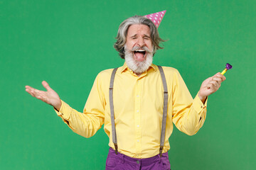 Crazy screaming elderly gray-haired mustache bearded man in yellow shirt suspenders birthday hat hold pipe spreading hands keeping eyes closed isolated on green colour background, studio portrait.