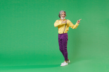 Fototapeta na wymiar Full length side view of excited elderly gray-haired mustache bearded man in casual yellow shirt suspenders pointing index fingers aside isolated on bright green colour background, studio portrait.