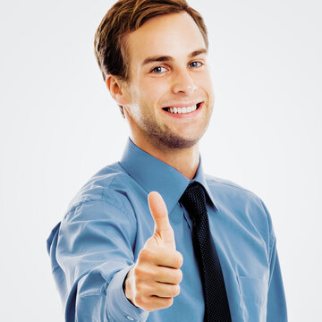 Portrait image - smiling businessman showing thumbs up like hand sign gesture, over grey background. Happy man in blue confident clothing, gesturing. Square composition.