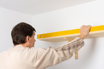 A man hangs a shelf in an apartment on a white wall, a worker checks the evenness of the shelf with a spint level
