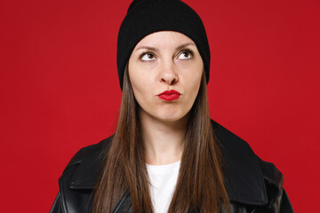 Close up of pensive puzzled beautiful young brunette woman 20s wearing basic casual black leather jacket white t-shirt hat looking aside up isolated on bright red colour background, studio portrait.