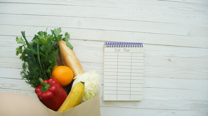 Full paper bag of food products with blank little notebook on wooden table, top view