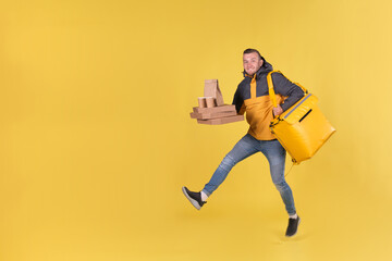 Fototapeta na wymiar Blond unshaven teen food delivery boy and courier in yellow clothes with thermo bag on his back holds an order for customer and runs to deliver order on time in 15 minutes. Food delivery in city