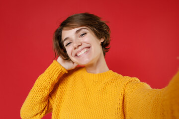 Close up of smiling attractive young brunette woman 20s in casual yellow sweater standing doing selfie shot on mobile phone put hand on head isolated on bright red colour background studio portrait.