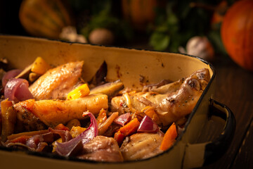 Baked chicken with vegetables in an iron brazier on a dark wooden background, chicken drumsticks with eggplant, pumpkin, onions and carrots baked in the oven