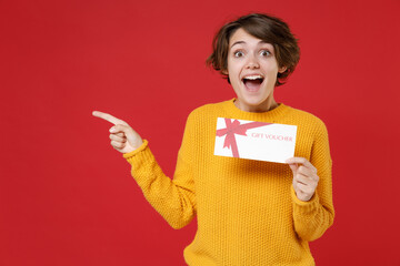 Surprised excited young brunette woman 20s wearing basic yellow sweater standing hold in hands gift certificate pointing index finger aside isolated on bright red colour background studio portrait.