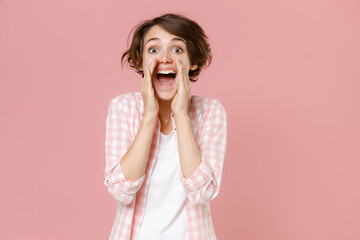 Excited cheerful young brunette woman 20s wearing casual checkered shirt standing screaming with hands gesture near mouth looking aside isolated on pastel pink colour background, studio portrait.