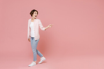 Fototapeta na wymiar Full length side view of excited shocked young brunette woman 20s wearing casual basic checkered shirt standing pointing index finger aside isolated on pastel pink colour background, studio portrait.