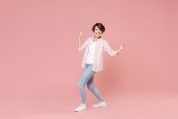 Fototapeta na wymiar Full length side view of overjoyed young brunette woman 20s wearing casual checkered shirt standing clenching fists doing winner gesture isolated on pastel pink colour background, studio portrait.