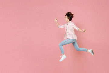 Fototapeta na wymiar Full length side view of smiling cheerful funny young brunette woman 20s wearing casual checkered shirt jumping like running clenching fists isolated on pastel pink colour background, studio portrait.