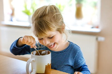 Adorable toddler girl drinking hot milk with froth. Happy healthy child indoors, enjoying chocolate drink.